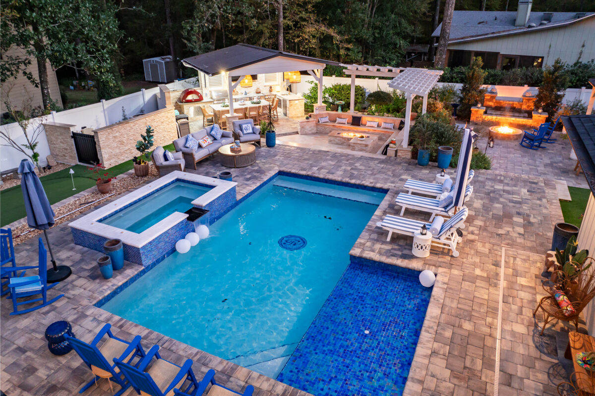 Aerial view of an entire hardscaped back yard, with custom pavers around the pool, a waterfall feature, two fire pits, and a custom outdoor kitchen